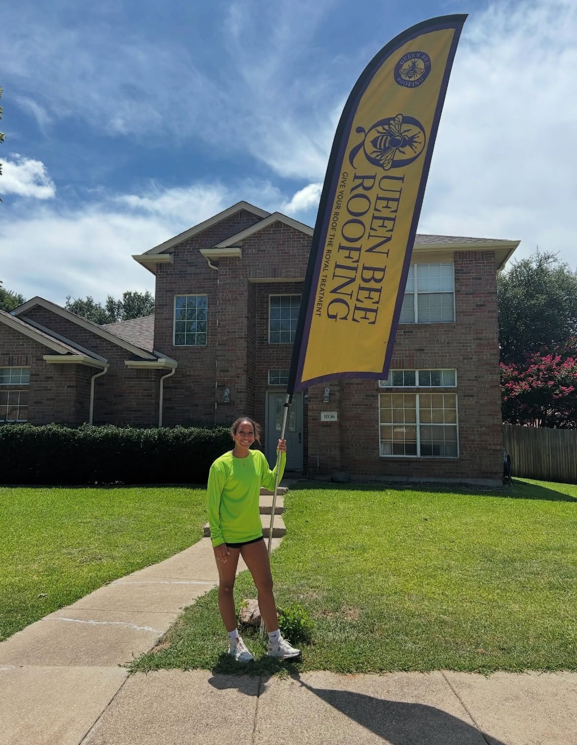 A woman holding a yellow "Queen Bee Roofing" feather flag in front of a two-story brick house